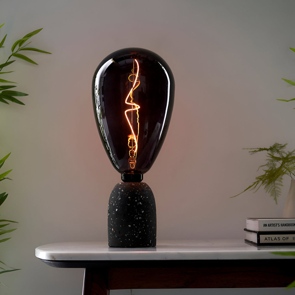 4W E27 LED Filament Wisp Bulb in smoke glass with warm white glow, teardrop shape on a black terrazzo lamp base atop a marble table.