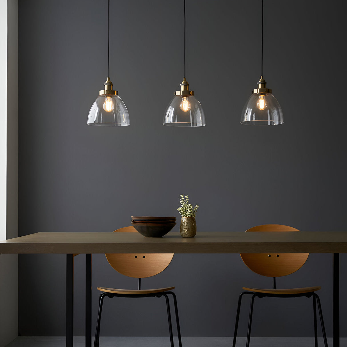 Wentworth 3 Light Linear Pendant in antique brass, clear glass dome shades with visible filament bulbs, hung over a dining table in a dark grey room.