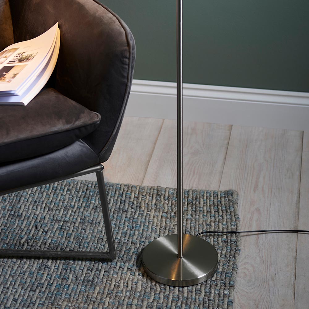Endon Brackbourne floor lamp base in satin nickel on a woven rug beside a dark velvet chair, with black flex trailing across pale wood flooring.