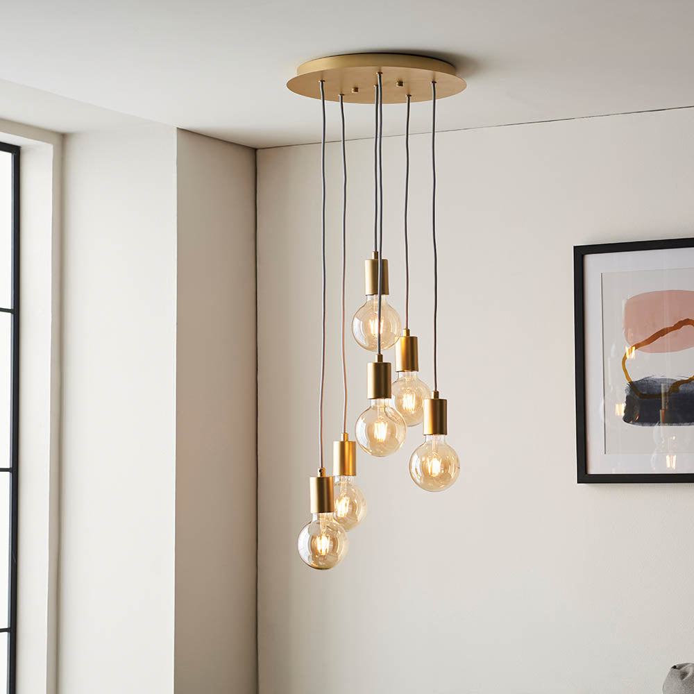 Marlowe 6 Light Pendant in gold, staggered exposed bulbs on grey flex cables from a round ceiling plate, shown in a room with framed wall art.