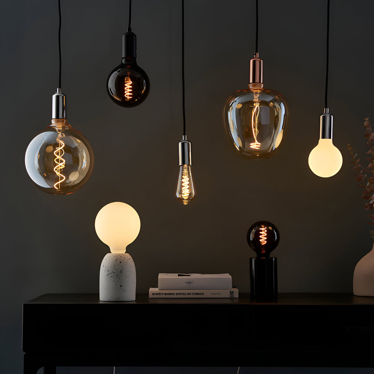 Endon amber LED filament pendant and table bulbs in varied shapes with warm glow, styled above a black console with books and foliage.