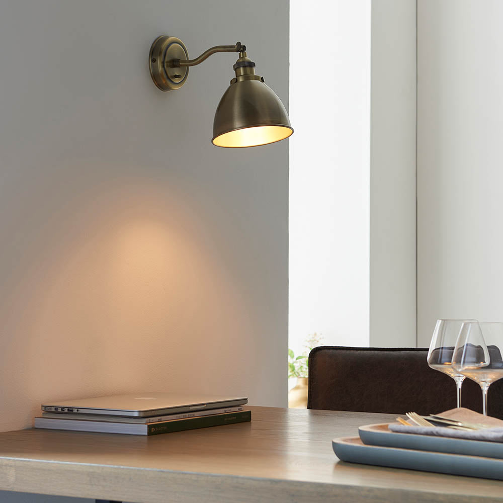 Endon Bramdale wall light in antique brass with adjustable knuckle and dome shade, mounted above a wooden dining table with laptop and wine glasses.