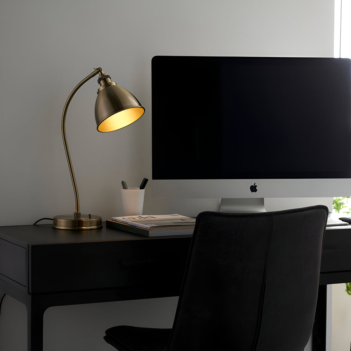 Endon Bramdale brass table lamp with curved arm and dome shade illuminated on a black desk beside an iMac in a modern home office.