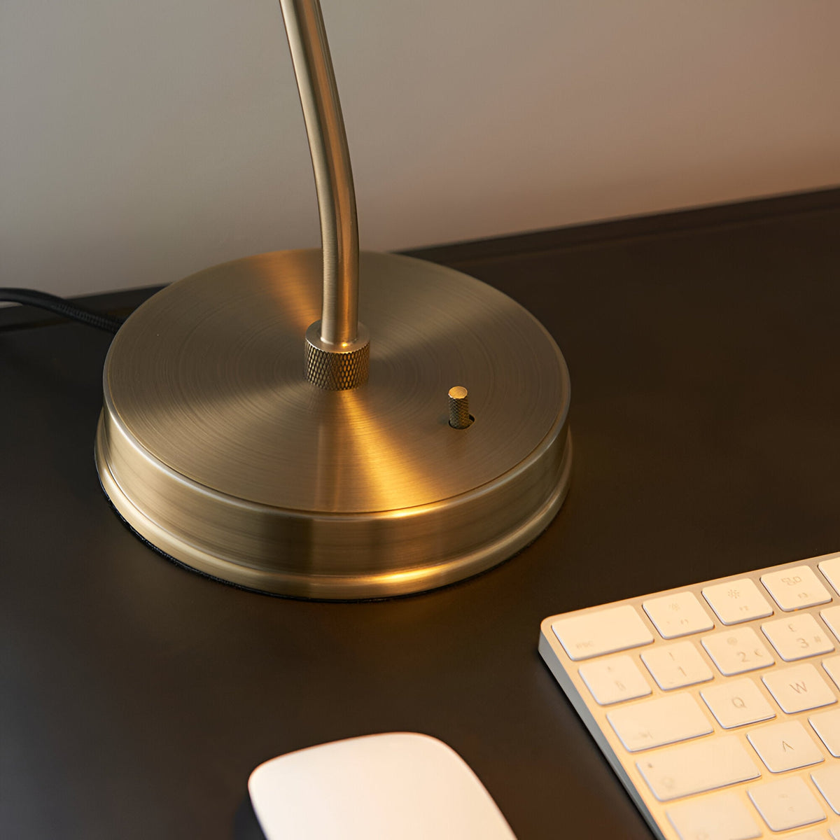 Close-up of the Endon Bramdale table lamp's round brushed brass base and toggle switch on a dark desk beside a keyboard and mouse.