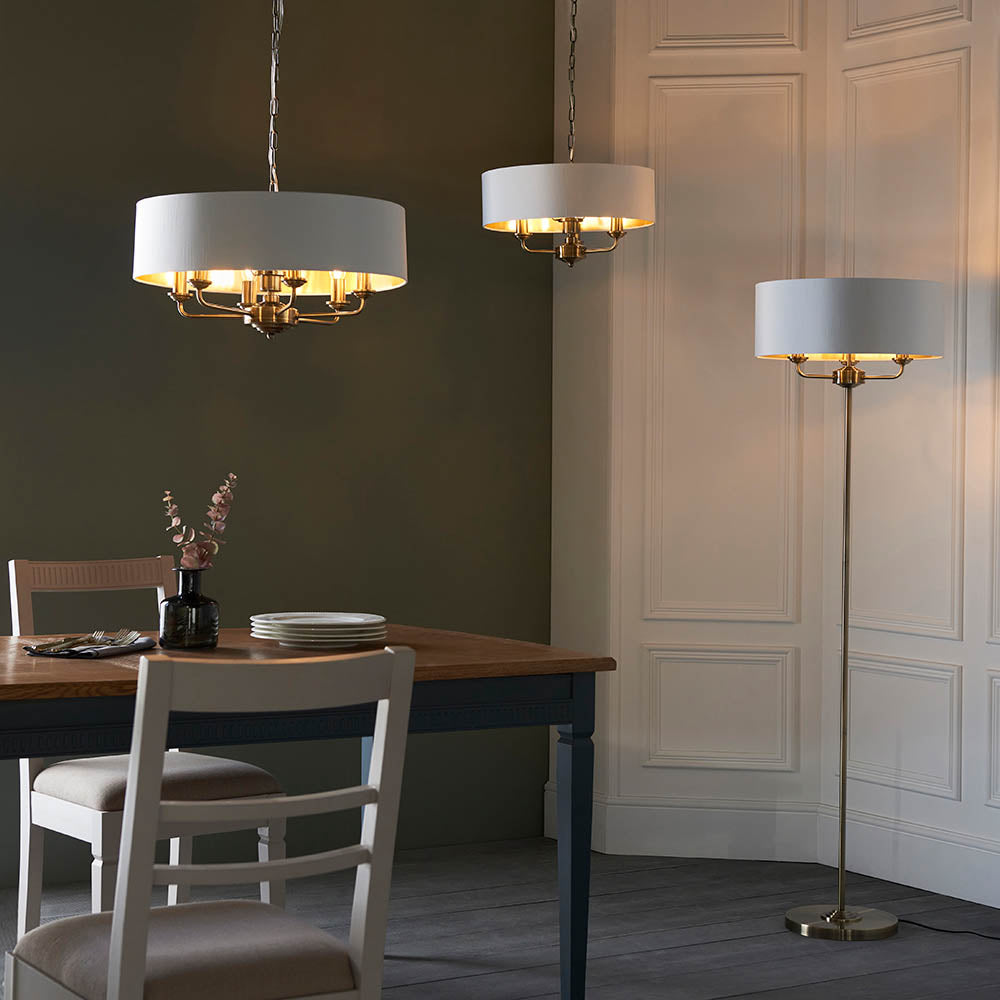 Endon Brenley V1 six-light pendants with white drum shades and brass fittings shown above a dining table beside a matching floor lamp in a panelled room.