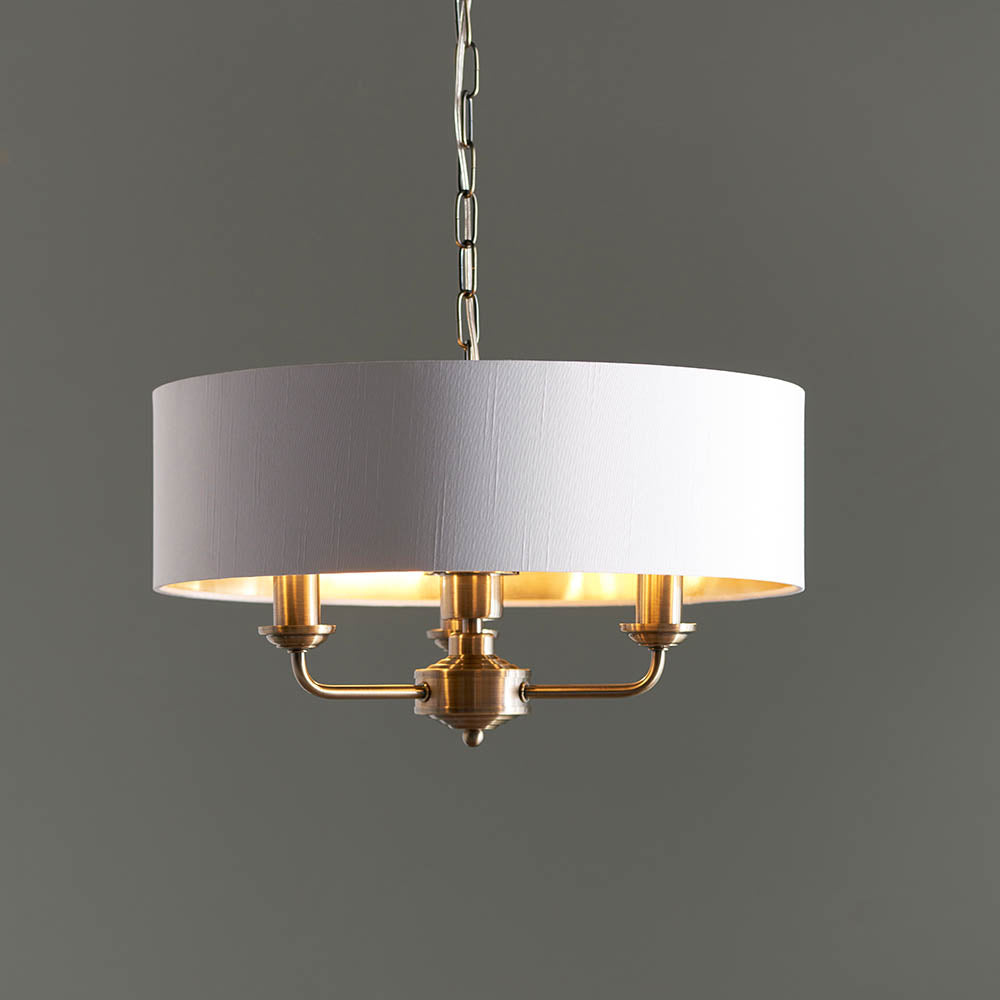 Endon Brenley 3 light pendant with a vintage white drum shade, gold metallic inner lining and antique brass fittings on a chain, against a grey background.