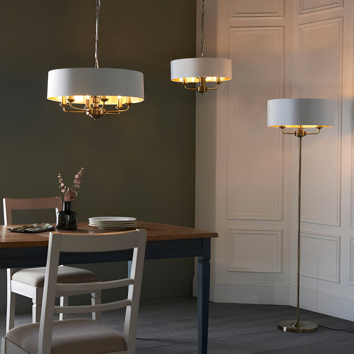 Endon Brenley 3 light floor lamp with white drum shade and brass frame, shown beside matching pendants over a dining table in a dark green room.