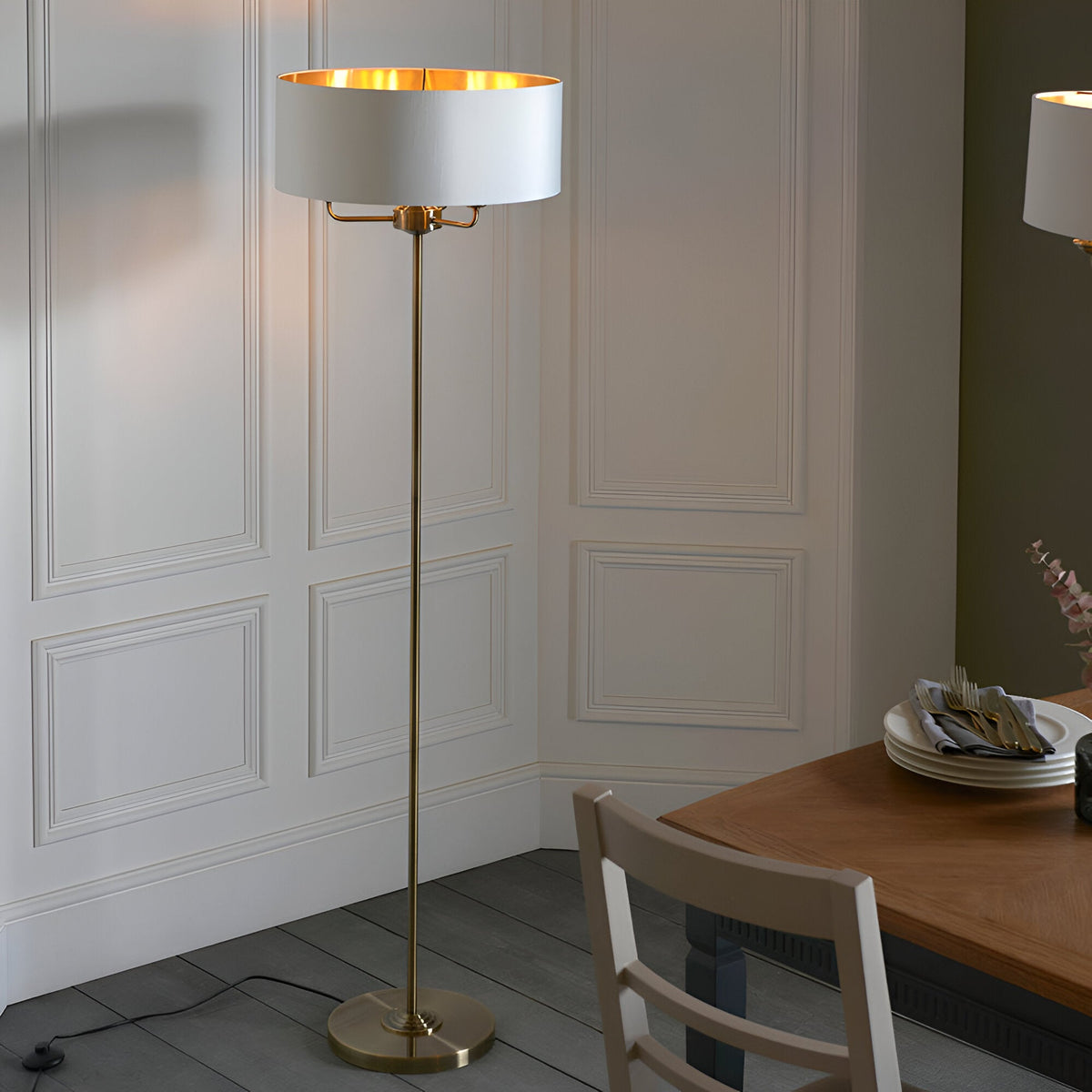 Endon Brenley 3 light floor lamp with brass stem and white drum shade lined in gold, shown beside a dining table against panelled walls.