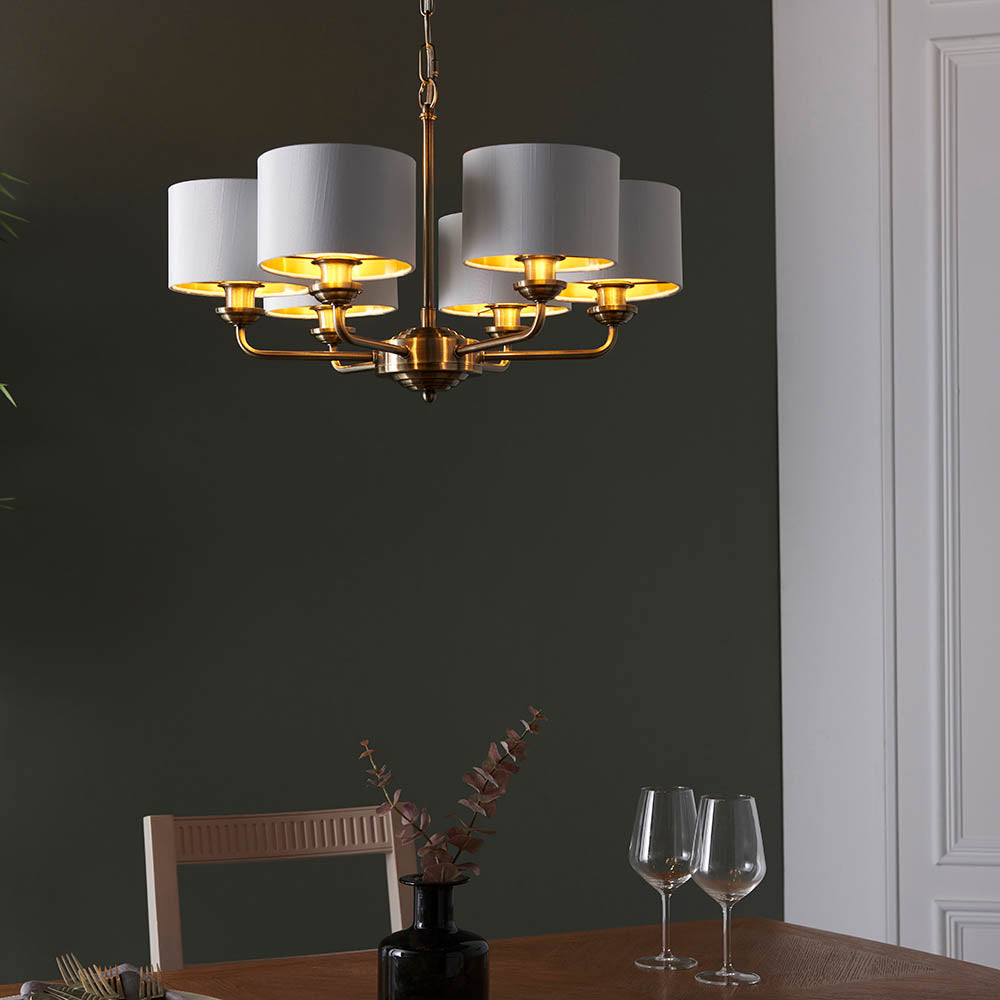 Brenley V2 6-light pendant with antique brass arms and white fabric drum shades, shown lit above a dining table in a dark green room.