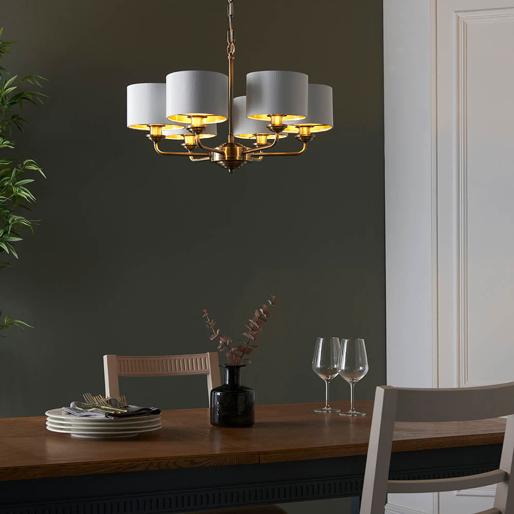 Brenley V2 six-light pendant with antique brass metalwork and white fabric drum shades, hung above a dining table in a dark-walled room.