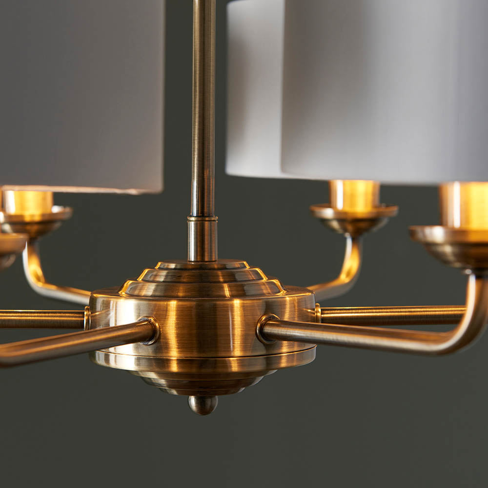Close-up of the Brenley V2 6 Light Pendant's antique brass centre column and multi-arm frame with white fabric shades, set against a dark background.
