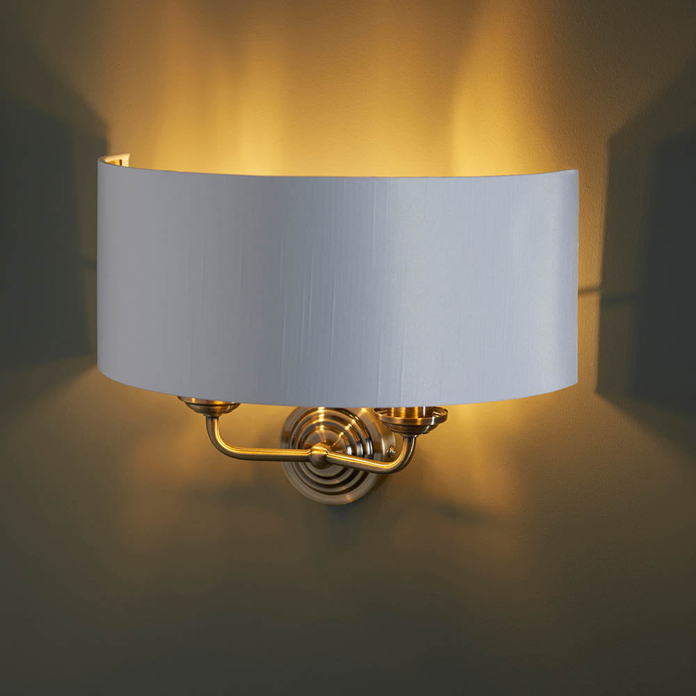 Endon Brenley 2 light wall light with a curved white linen shade on an antique brass backplate, casting a warm upward glow against a neutral wall.