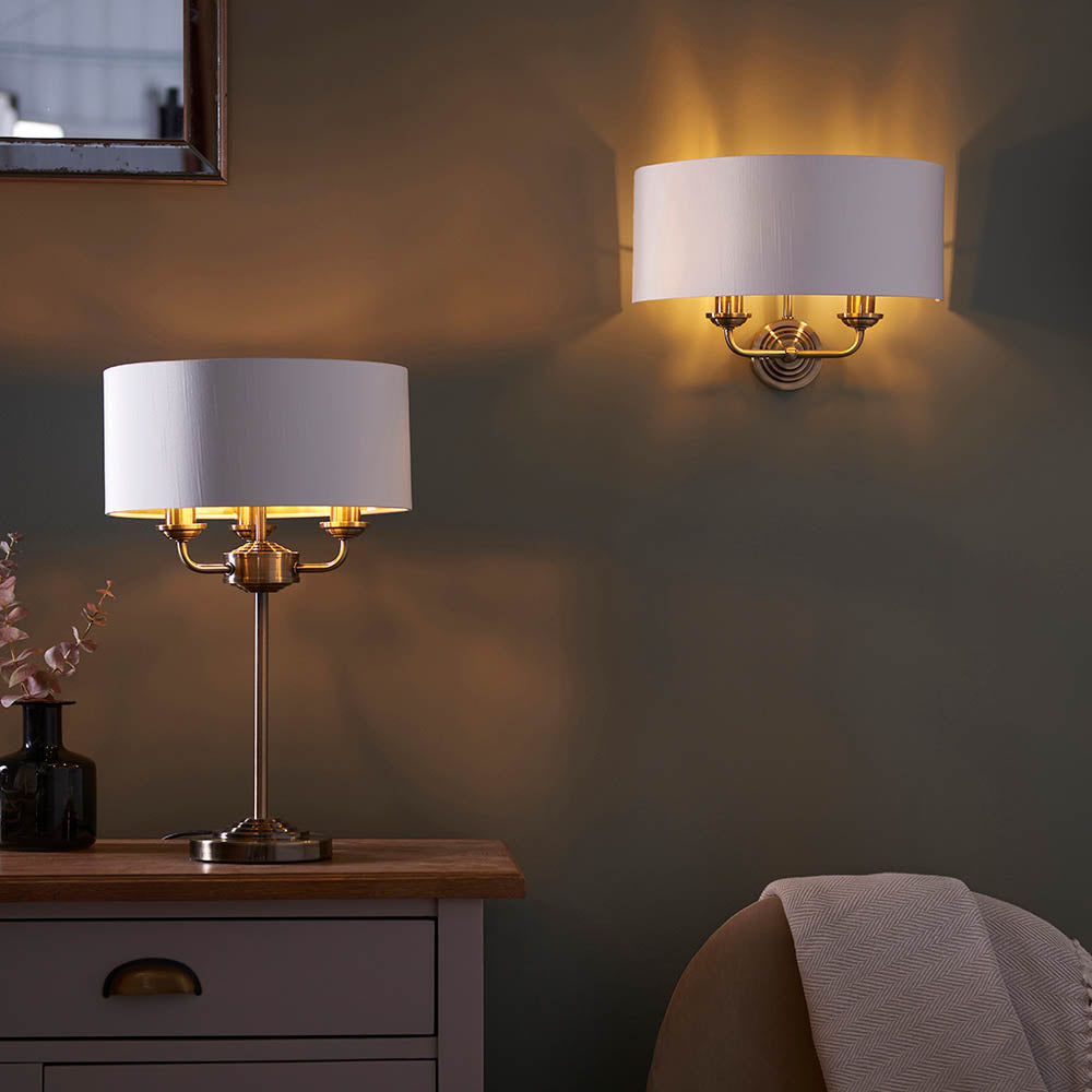 Endon Brenley 2 Light Wall Light in brass and gold with a white linen half-shade, illuminated above a table lamp on a sideboard in a dim living room.