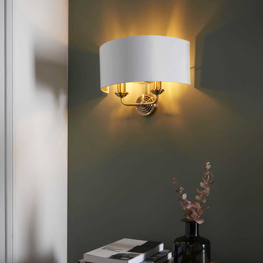 Endon Brenley 2 light wall light with white linen half-shade and antique brass twin arms, mounted on a green wall above a vase of eucalyptus.