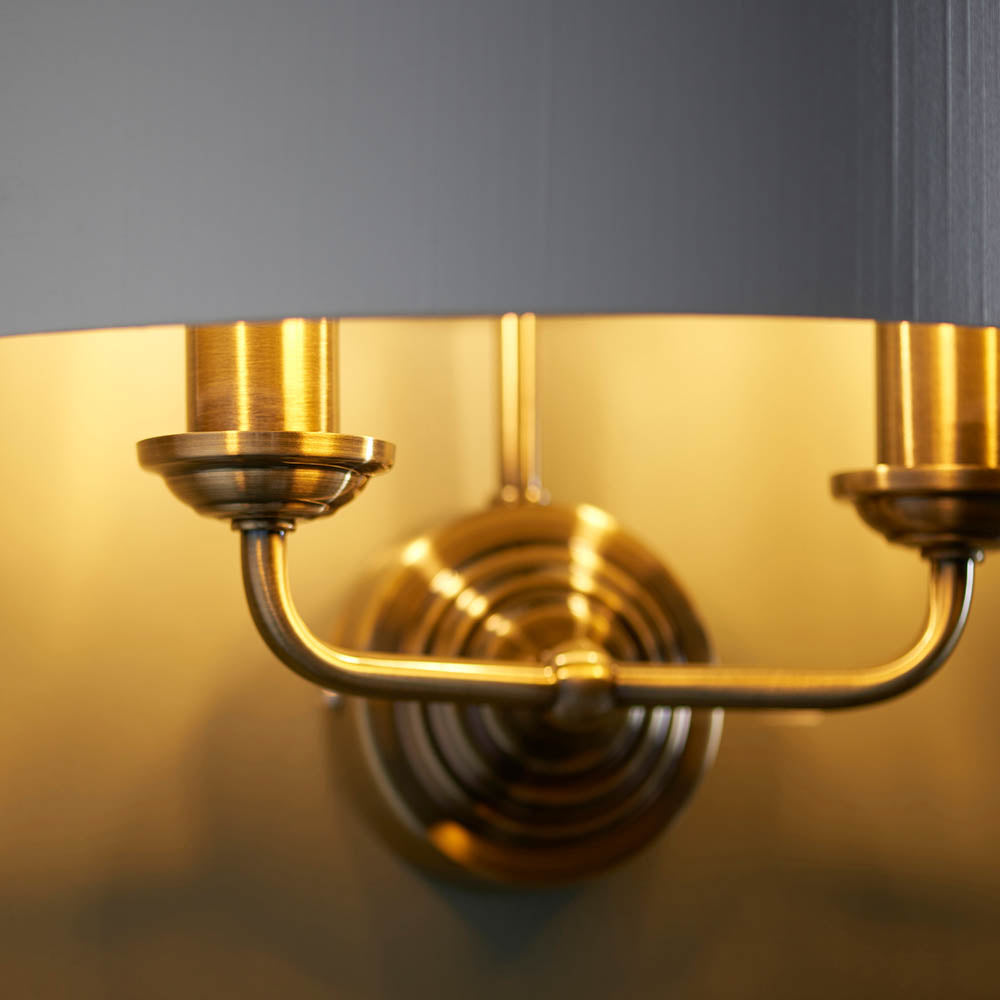 Close-up of Endon Brenley 2 Light Wall Light showing antique brass twin arms, ribbed backplate and gold-lined shade interior against a grey wall.