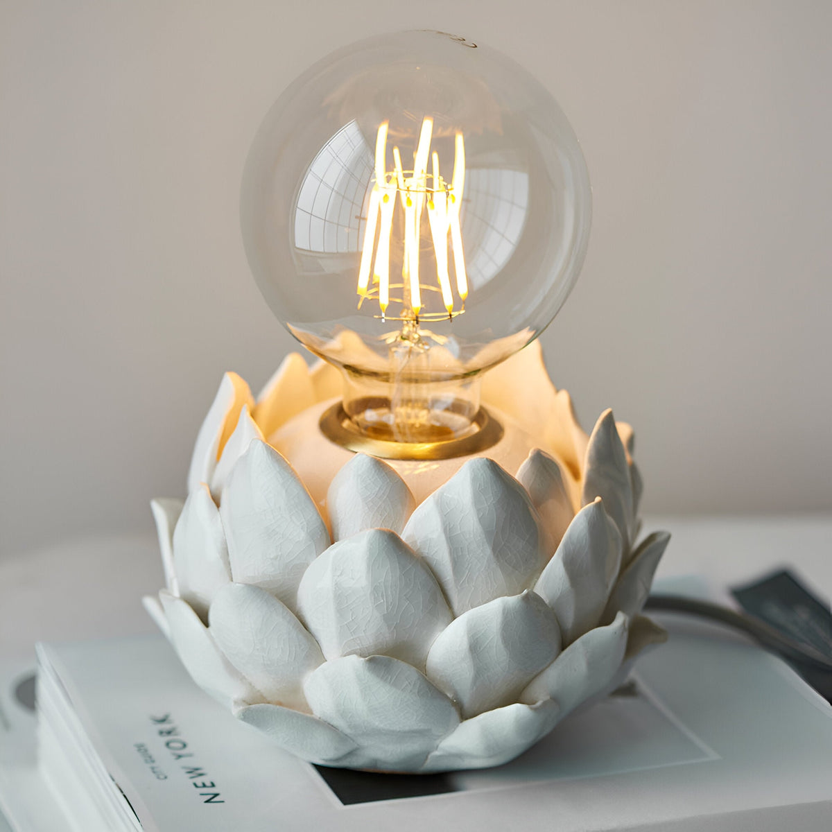 Aldgrove Table Lamp with white ceramic artichoke-style layered petal base, antique brass neck detail, and lit E27 globe bulb, shown on a book.