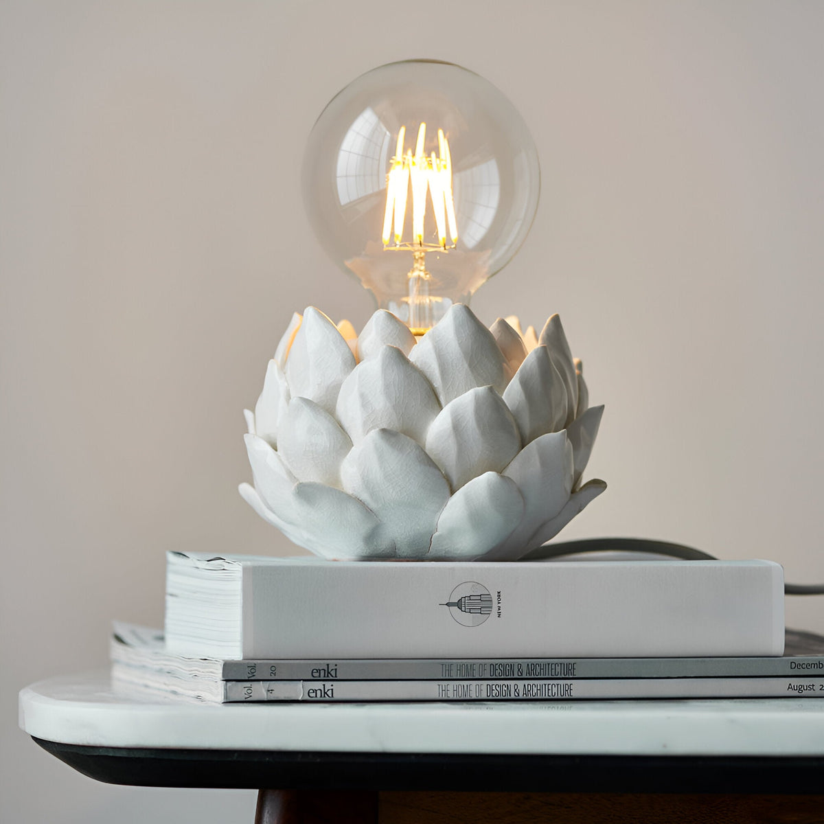 Aldgrove Table Lamp in white ceramic artichoke design with antique brass neck detail, shown lit with a clear E27 bulb atop stacked books.