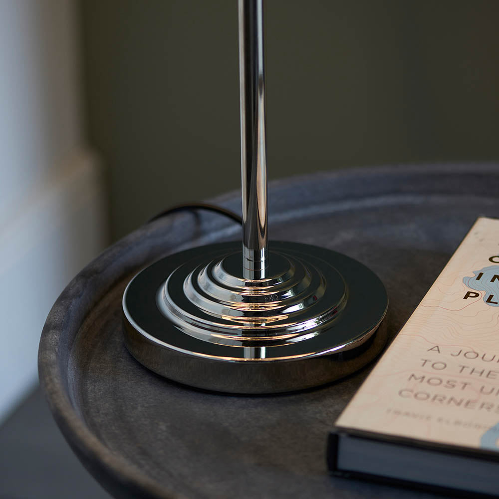 Close-up of the Endon Brenley 3 Light Table Lamp's tiered nickel base and stem, resting on a dark wooden side table beside a book.
