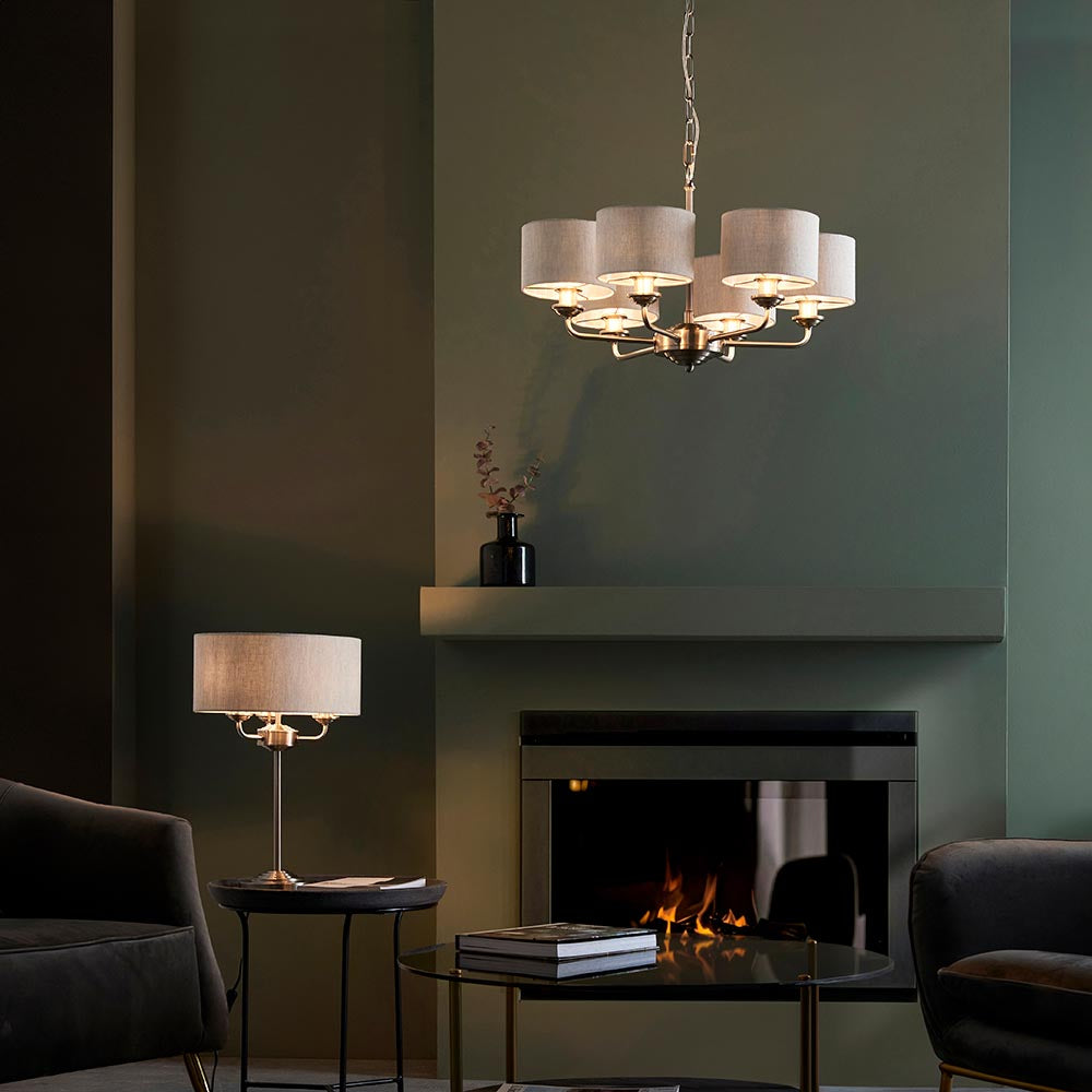 Brenley 6 Light Pendant in brass and chrome with fabric drum shades, shown above a fireplace in a living room, paired with a matching table lamp.