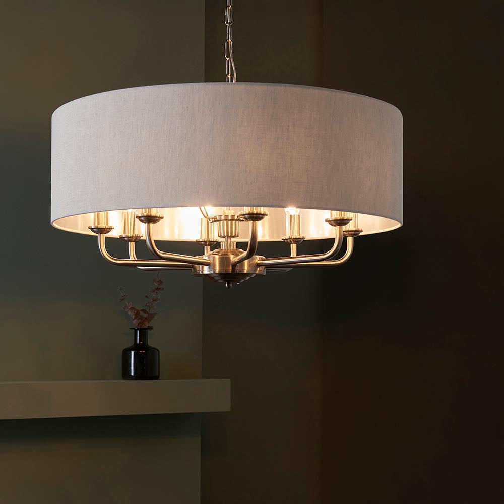 Endon Brenley 8 light pendant in chrome and brass with a large drum shade in natural linen, hanging in a dimly lit room above a shelf.