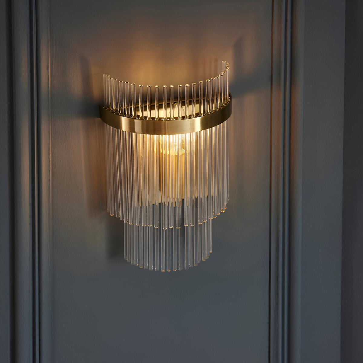 Beaumont Wall Light in brass, tiered clear glass rods surrounding a lit bulb on a round brass backplate, mounted on a grey panelled wall.