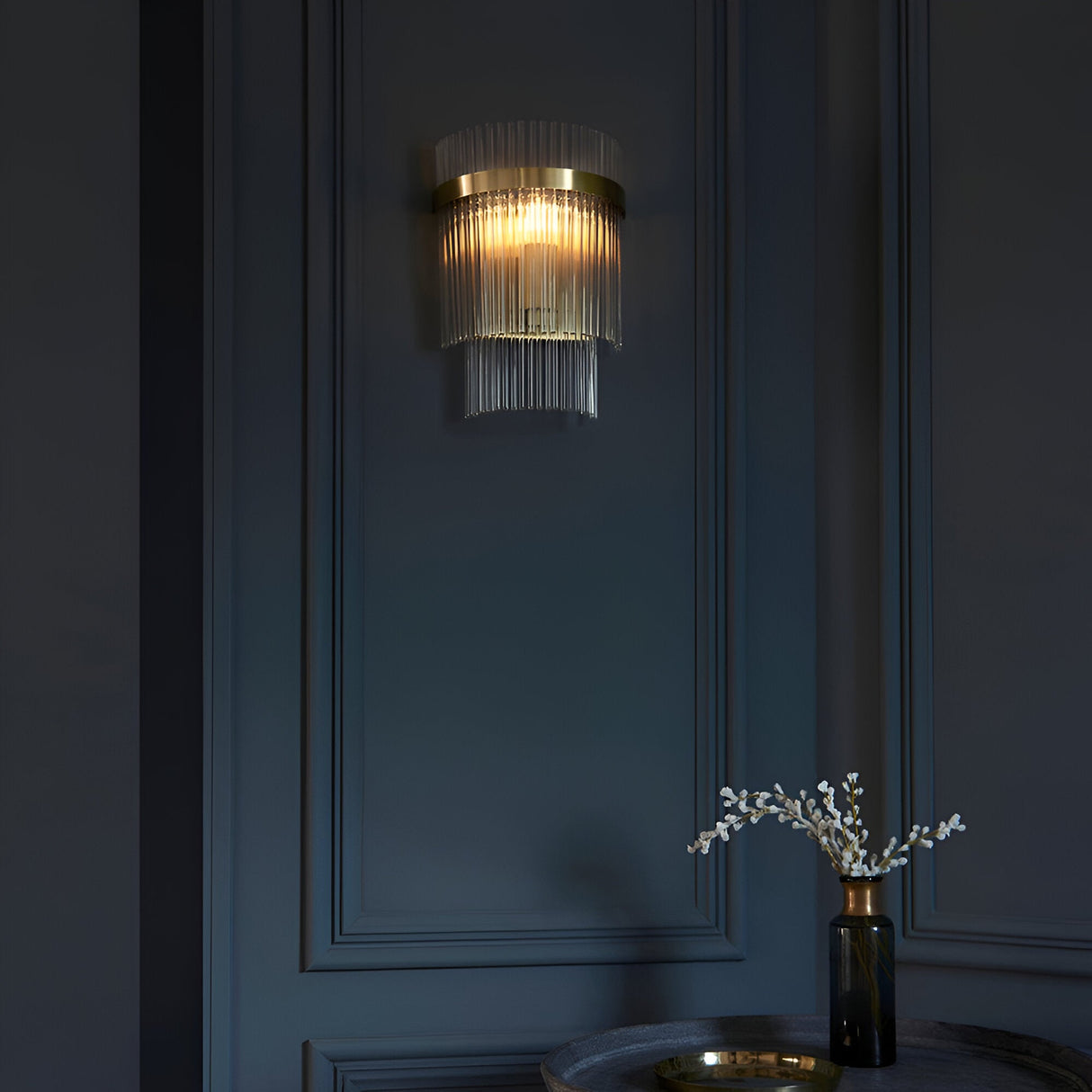 Beaumont Wall Light in brass with clear glass rods, glowing warmly on a dark panelled wall beside a table with a small vase.