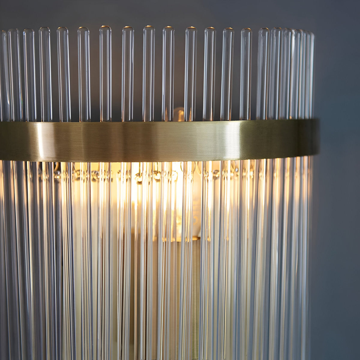 Beaumont Wall Light in brass, close-up of clear glass rods lit from within, with a brushed brass band and backplate against a grey background.