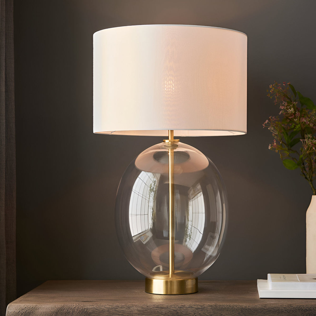 Brockmere Table Lamp in Brass with oval clear glass base and white fabric drum shade, shown lit on a wooden table beside a vase of flowers.