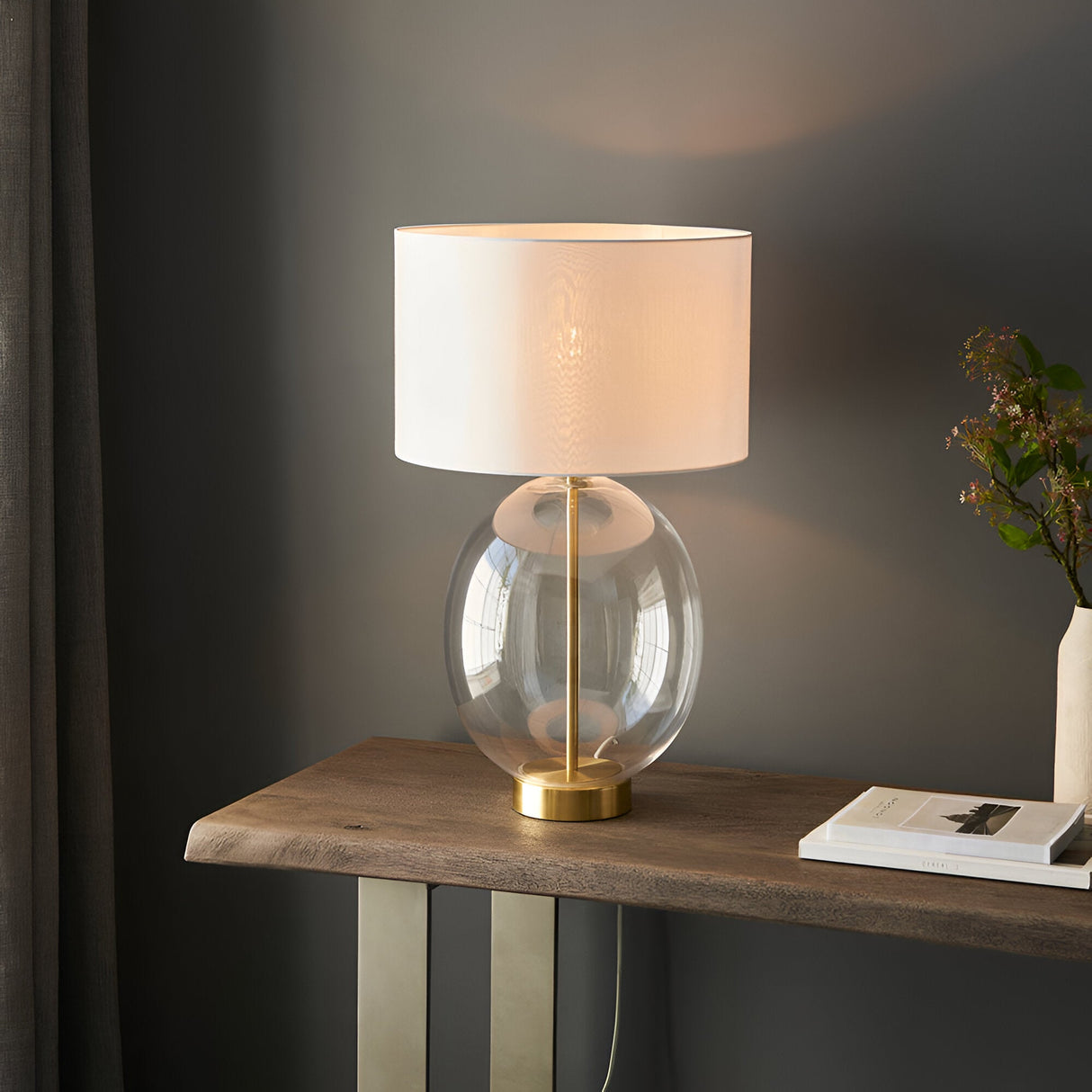 Brockmere Table Lamp in brass with an oversized oval clear glass base and white fabric drum shade, shown lit on a wooden console table.