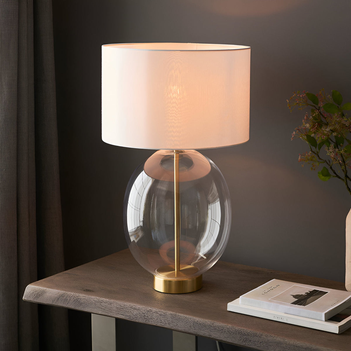 Brockmere Table Lamp in Brass with oval clear glass base and white fabric drum shade, shown lit on a console table beside books and foliage.
