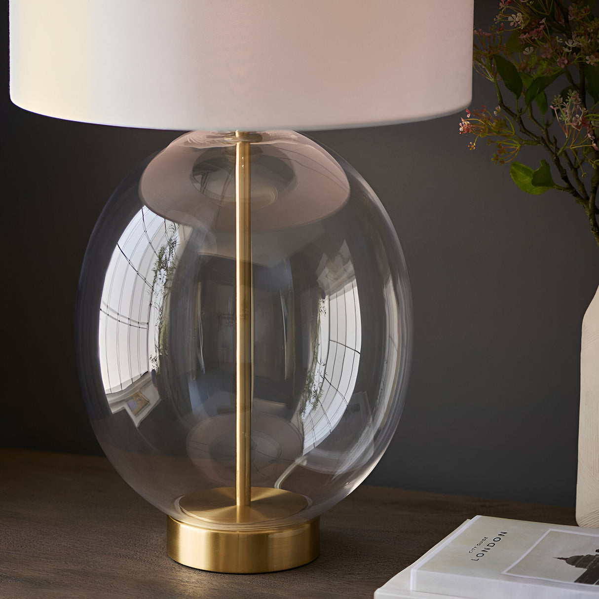 Brockmere Table Lamp with oversized oval clear glass base, satin brass stem and fitting, and white fabric shade, shown on a wooden surface.