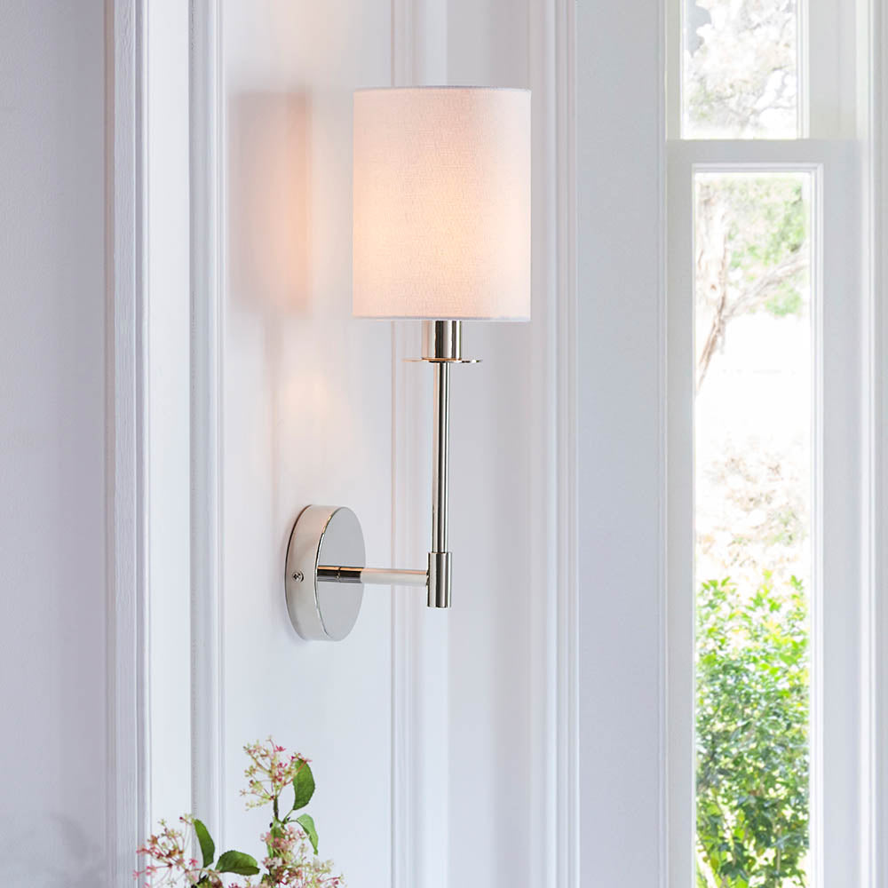 Endon Cliffhurst wall light with a slim polished nickel arm and vintage white cylinder shade, mounted on a panelled wall beside a bright window.