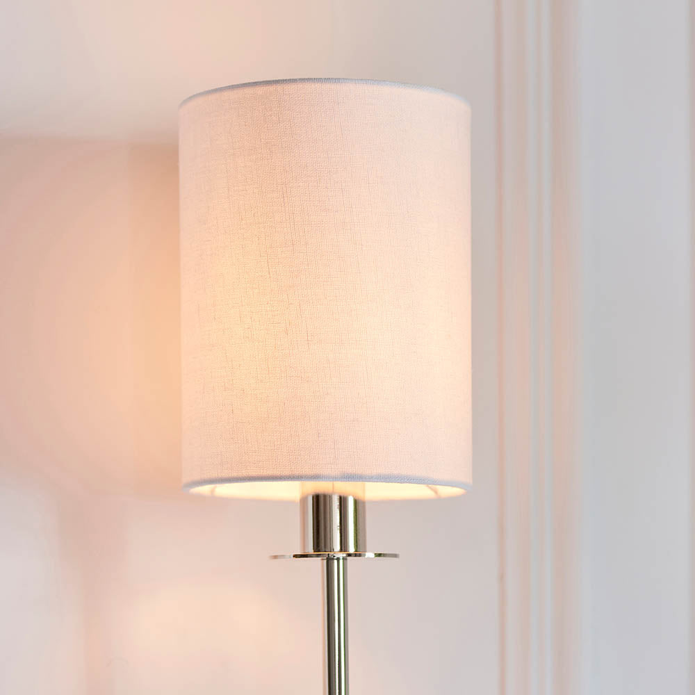 Endon Cliffhurst wall light illuminated on a panelled wall, featuring a slim polished nickel upright and a vintage white cylinder fabric shade.