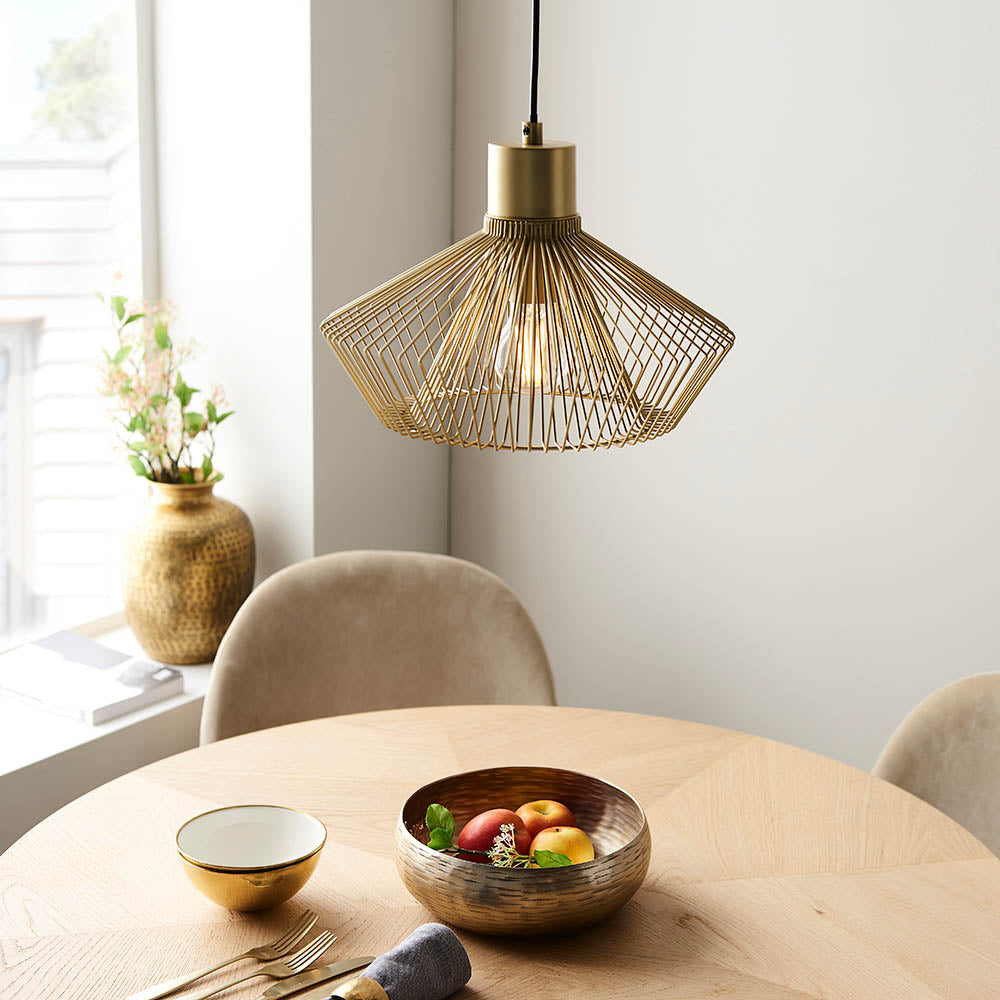 Endon Brockton cage pendant light in polished gold, hanging over a round wooden dining table set with a fruit bowl and beige velvet chairs.