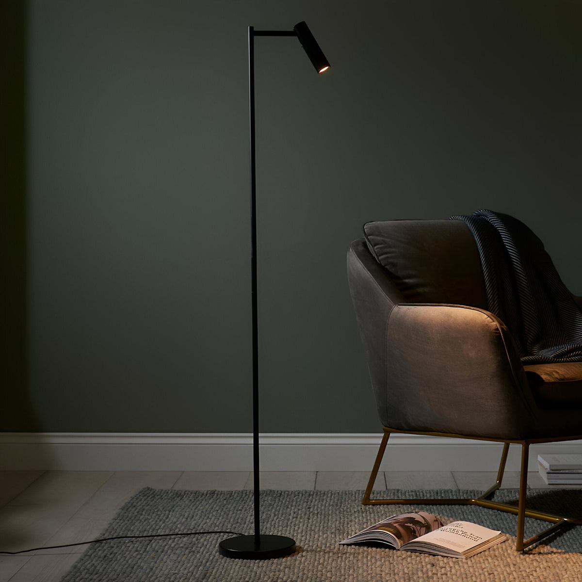 Endon Brackbourne matt black LED floor lamp with adjustable cylindrical head and round base, positioned beside a grey velvet armchair in a dim living room.