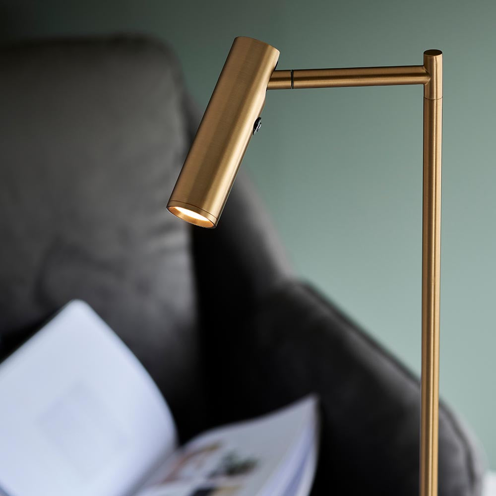 Endon Brackbourne brass floor lamp with adjustable cylindrical head and push button switch, angled over a dark armchair with an open book against a green wall.