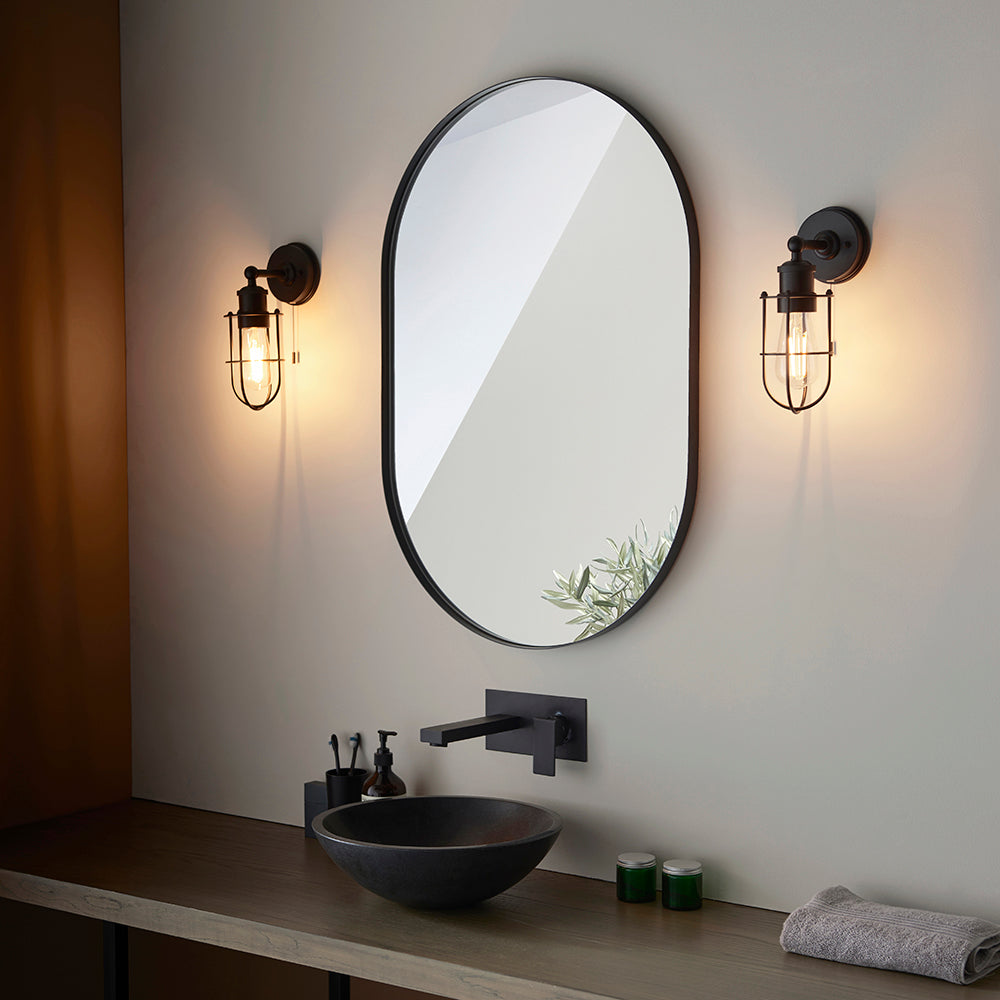 Bevwood black bathroom wall lights flank an oval mirror above a vanity, featuring industrial cage design, filament bulb, and pull cord.