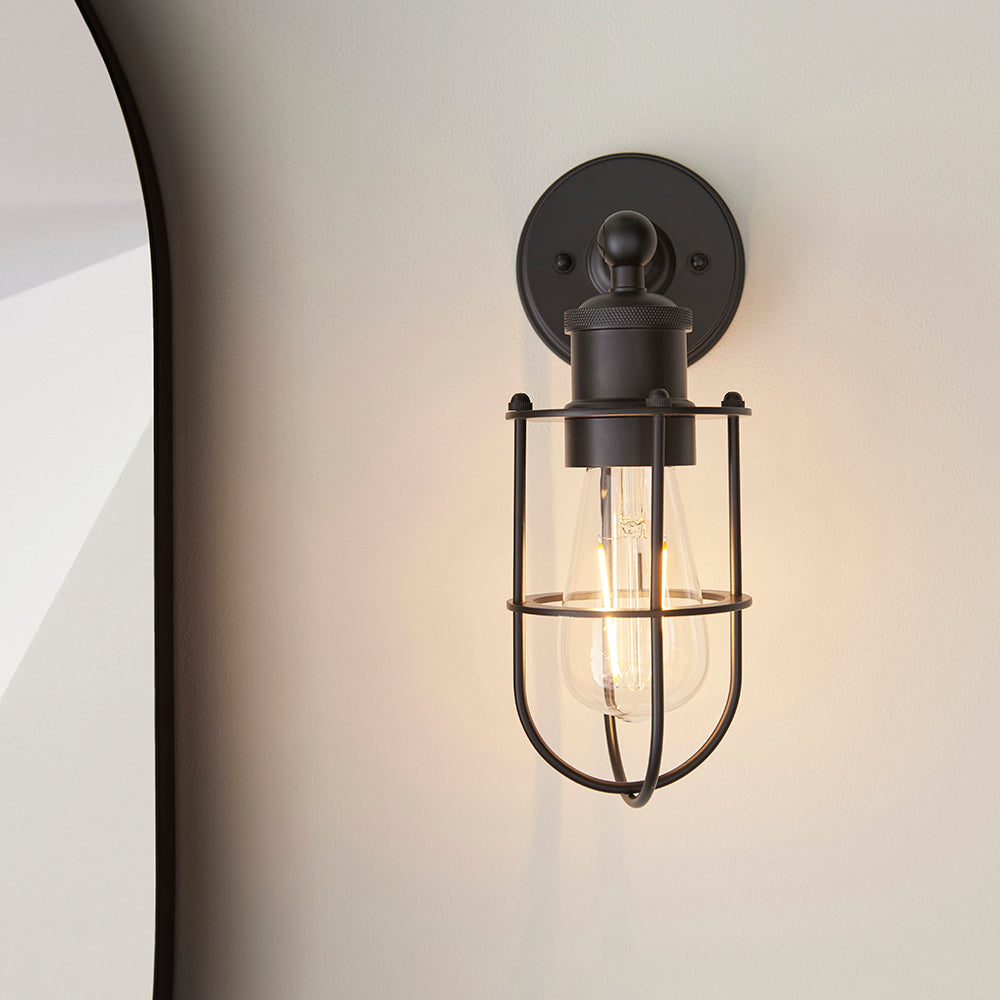 Bevwood industrial-style wall light in black metal with an open cage design and E27 filament bulb, mounted on a light neutral wall.