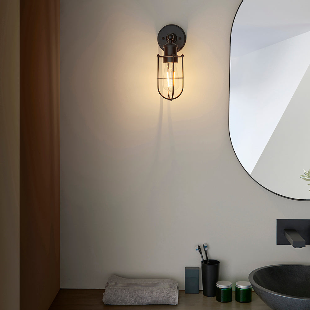 Bevwood industrial-style black wall light with cage design and visible filament bulb, mounted beside a mirror in a bathroom setting.