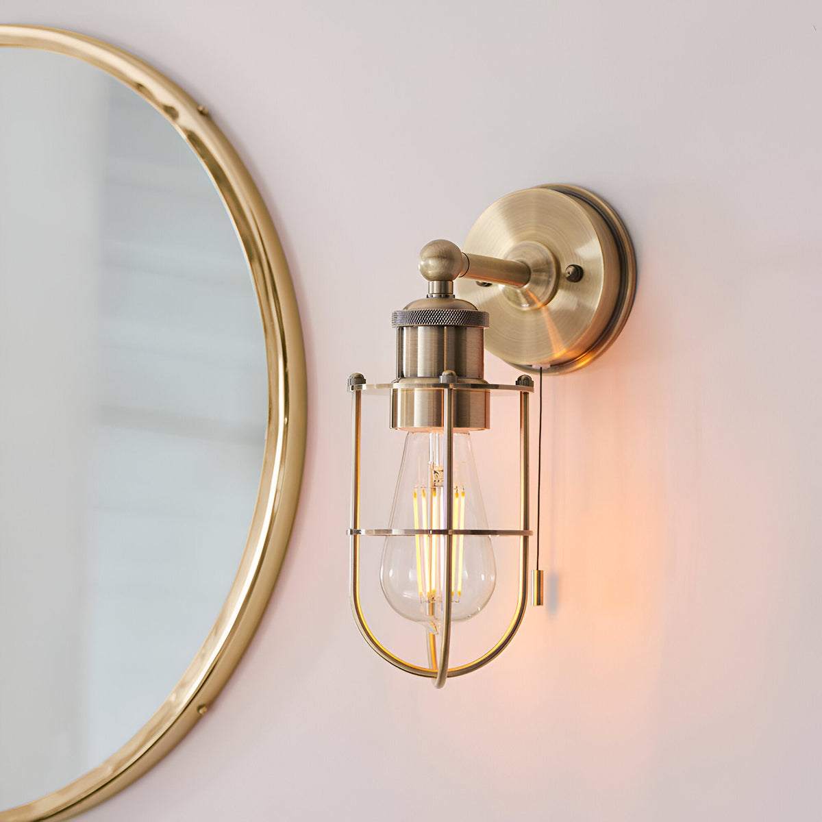 Bevwood industrial-style brass wall light with open cage design, exposed filament bulb and pull cord, mounted beside a round mirror.