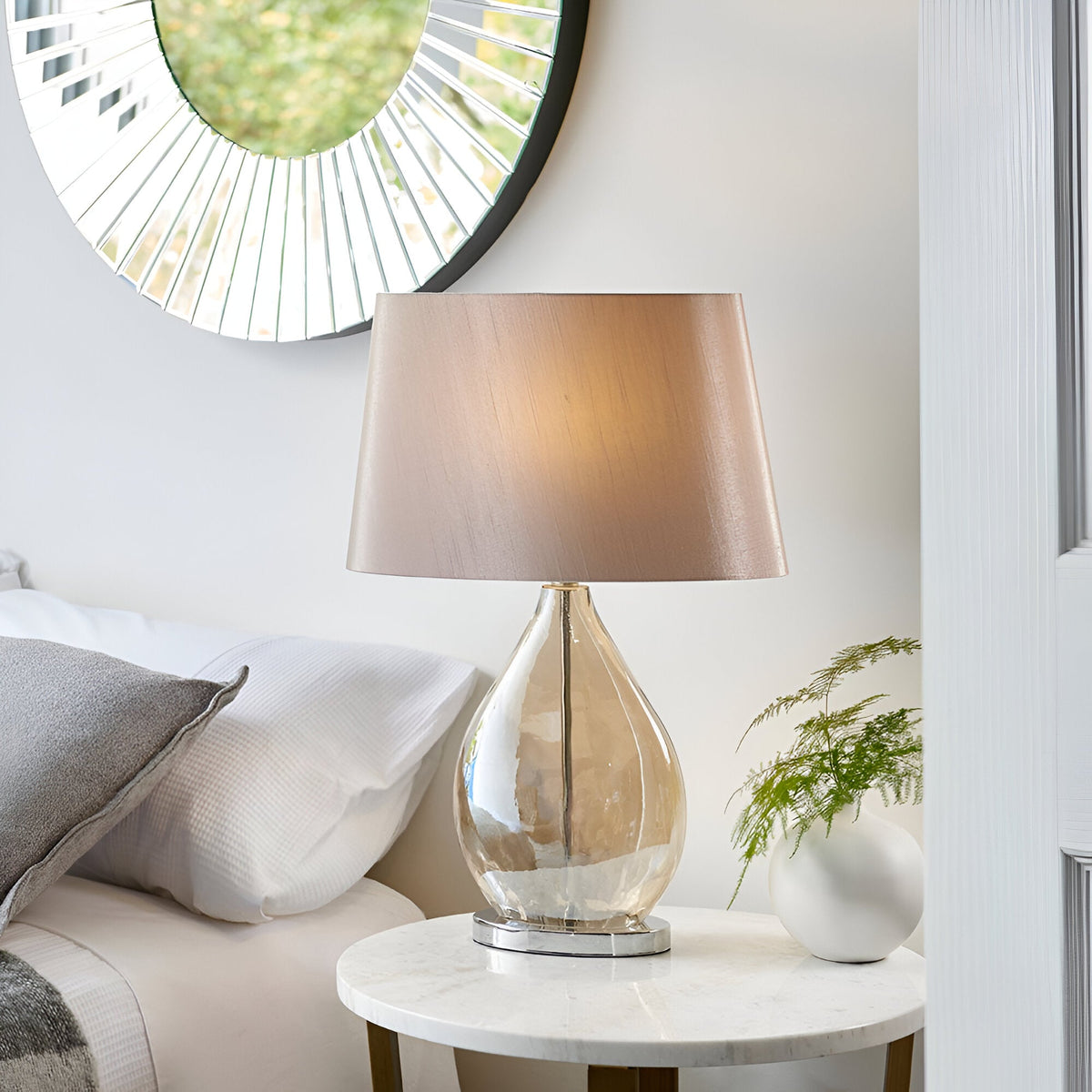 Brockstead Table Lamp in a bedroom setting, with a gold dimpled glass teardrop base, chrome fittings, and a mink oval faux silk shade on a marble side table.