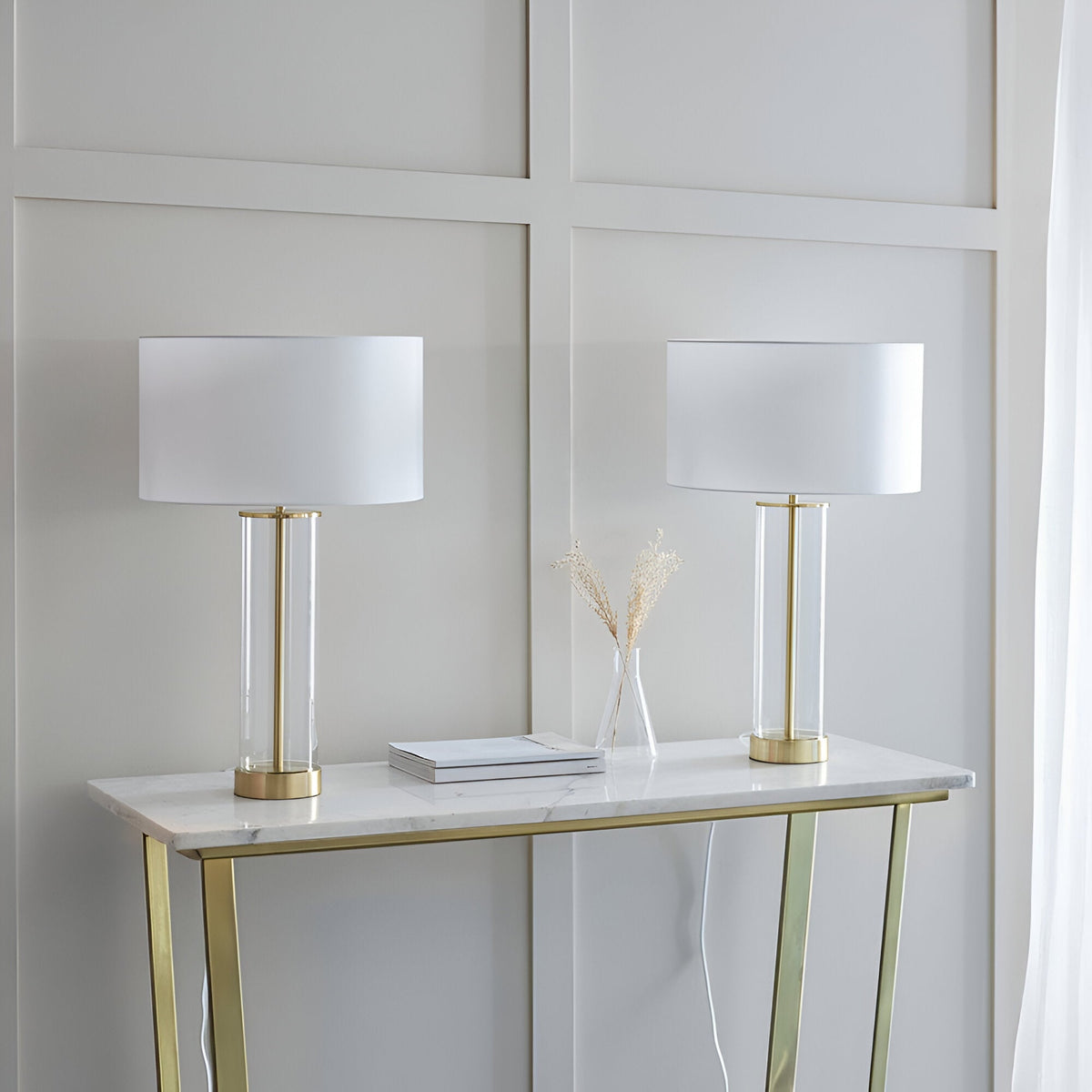 Endon Caldley twin pack table lamps with clear glass cylindrical bases, brass fittings and white drum shades, styled on a marble and gold console table.