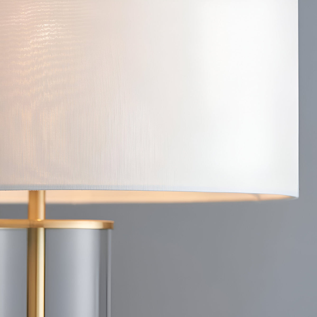 Close-up of the Endon Caldley table lamp showing its white faux silk drum shade, brass fitting and clear cylindrical glass base against a grey backdrop.