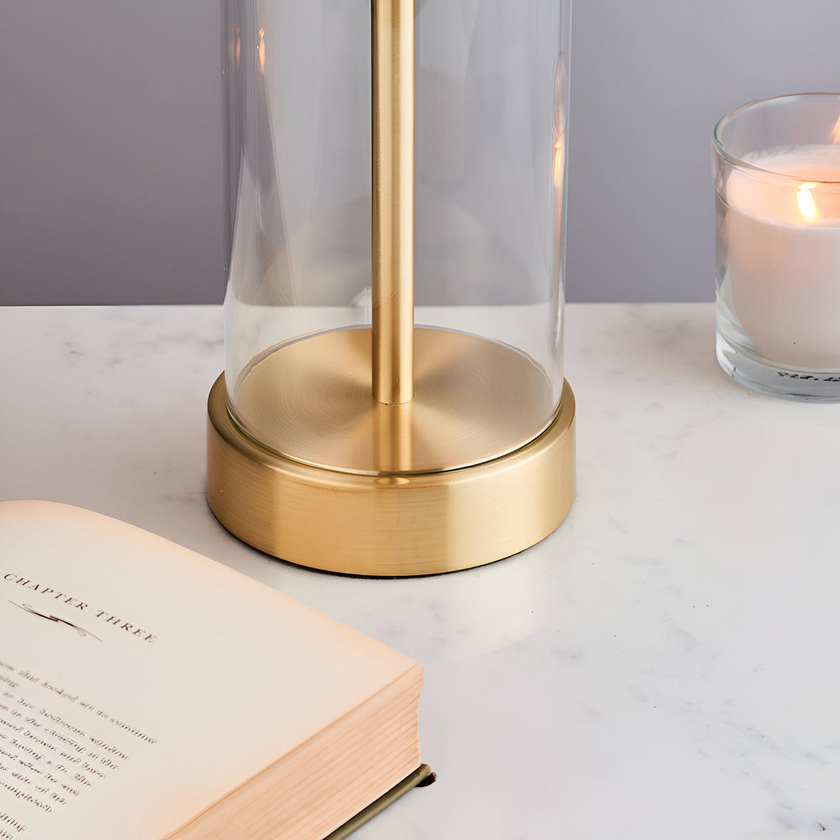 Close-up of Endon Caldley table lamp brass base with clear cylindrical glass, on a marble surface beside an open book and lit candle.