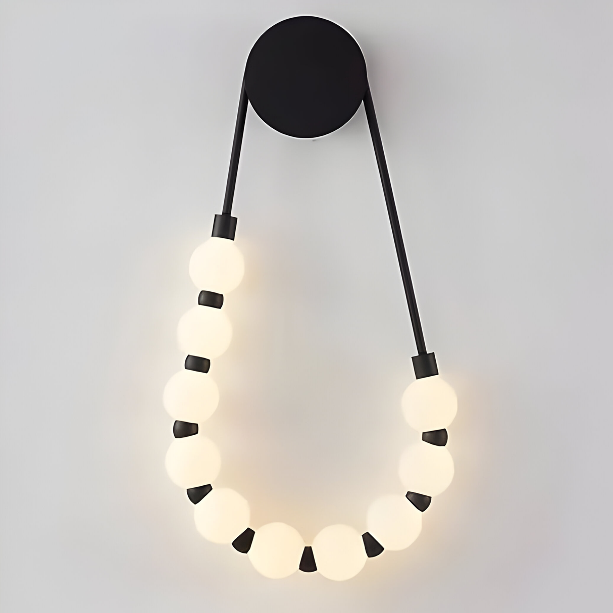 The Elixara 10 Light LED Alabaster Wall Light – White features a black circular base with two rods holding glowing alabaster bulbs in a bead-like loop, casting elegant ambient light on a light gray wall.