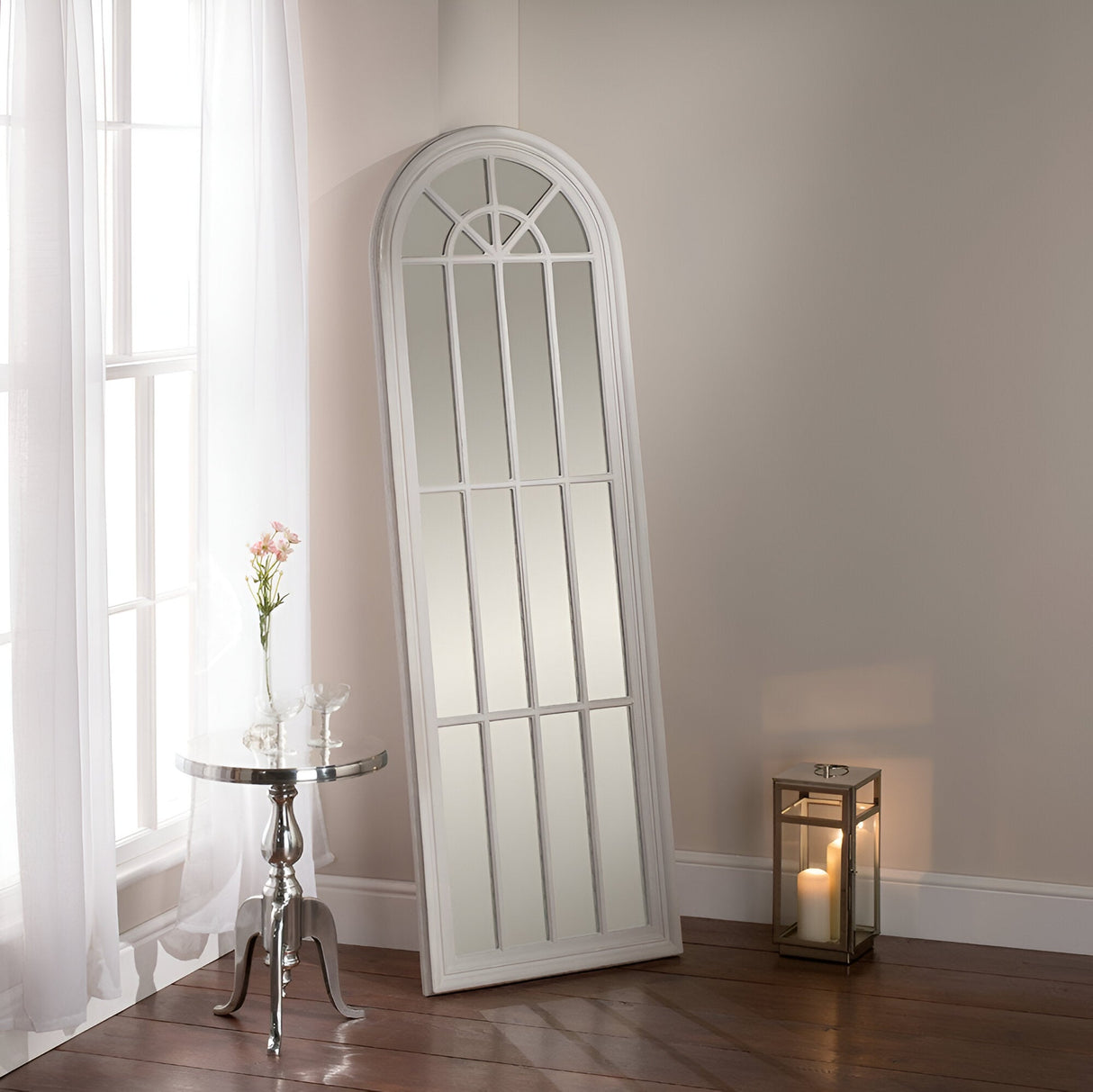 The Marquelle Arched Leaner Mirror in grey leans elegantly against a beige wall. Beside it, a round table displays a pink flower in a vase and two glasses. A candlelit lantern sits on the floor, as sunlight streams through sheer curtains.