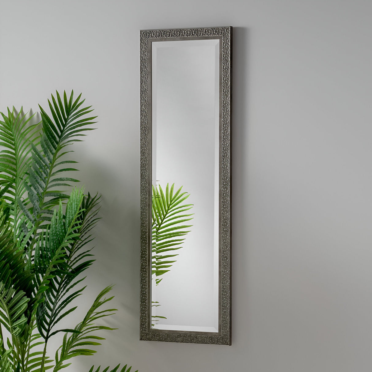 The Abbey Rectangle Wall Mirror in Grey, featuring its ornate frame, is installed on a plain wall, capturing the essence of minimalist design. Green palm fronds extend upward in front of it, their lush reflection adding a touch of elegance to the home decor.