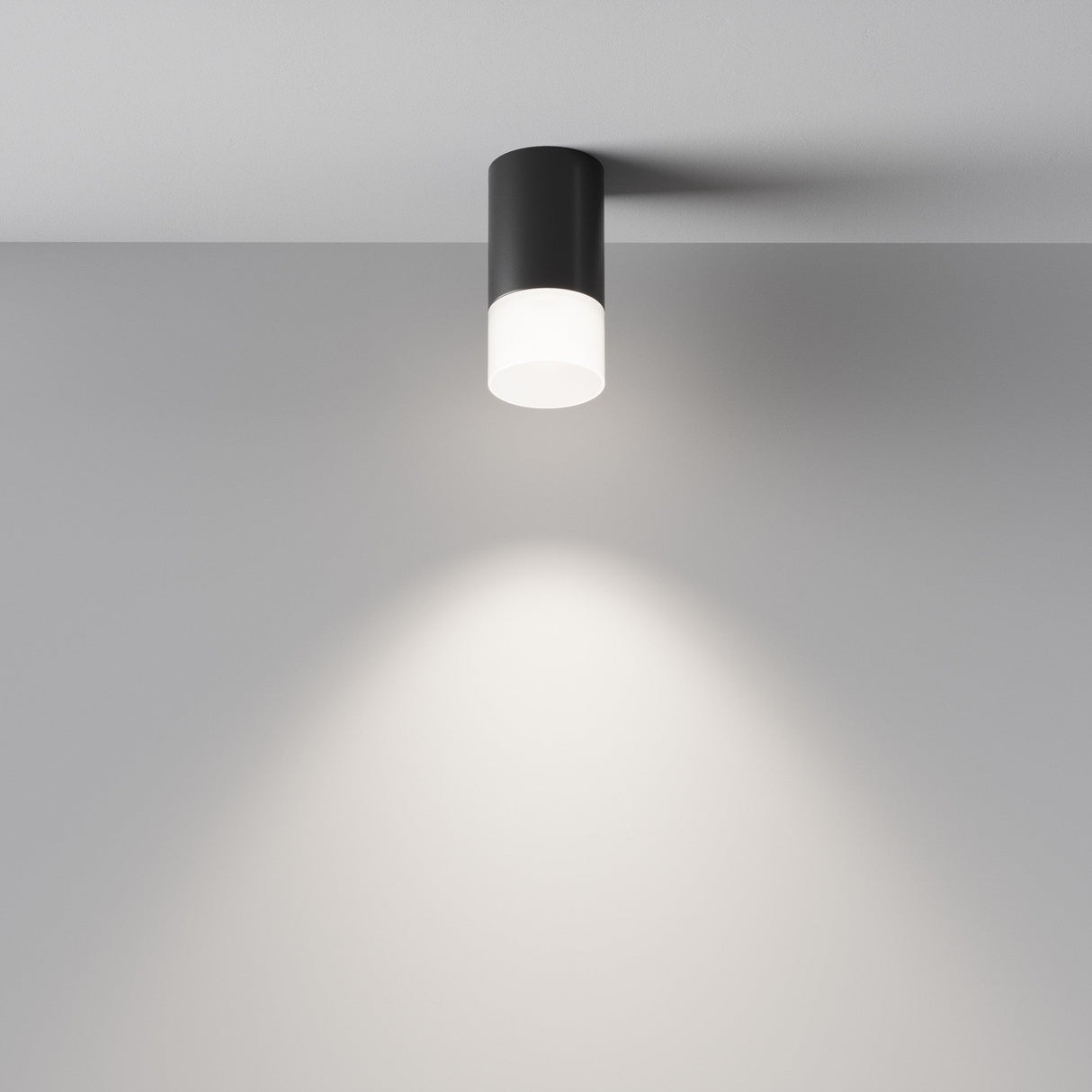 The Pauline Long Round Surface Downlight in matte black casts a soft, circular glow on a white wall. With its sleek, cylindrical design and frosted glass shade, it enhances ambient lighting with modern minimalism.