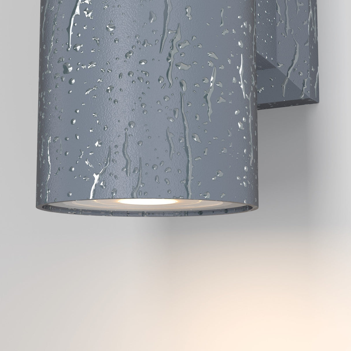 The Bowery Outdoor 2 Light Wall Light - Grey (O574WL-02GR) is shown close-up on a wall, shimmering with water droplets. Its durable cylindrical design emits a soft bidirectional glow, highlighting the textured, wet appearance of the surrounding wall.