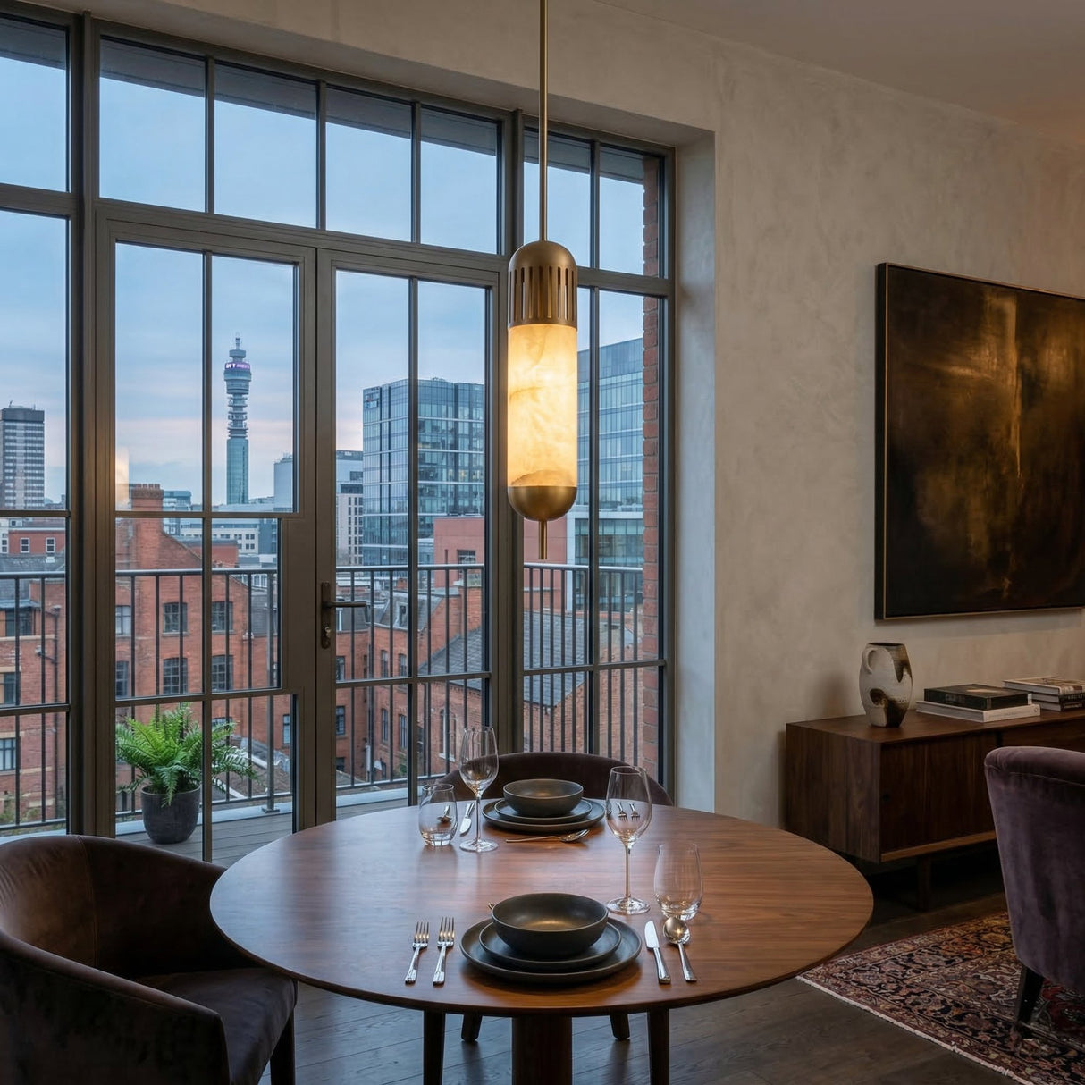 A round dining table for two with black plates and wine glasses sits by large windows overlooking the city, while the Zerith LED Fluted Tube Alabaster Single Pendant Light in soft white & brushed brass adds modern elegance overhead.